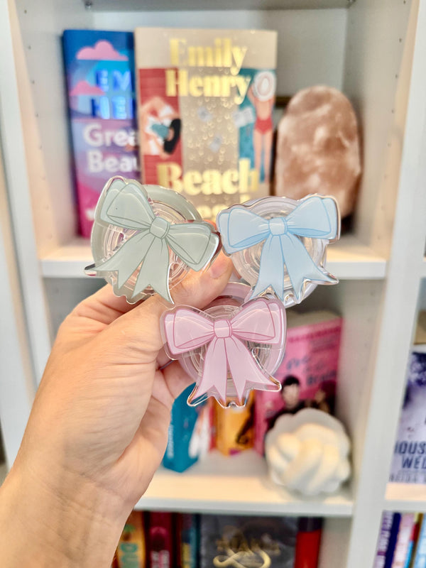 May include: Three clear acrylic bow-shaped phone grips in pastel shades: mint green, light blue, and pink. The grips are held in front of a bookshelf with books and a decorative item.