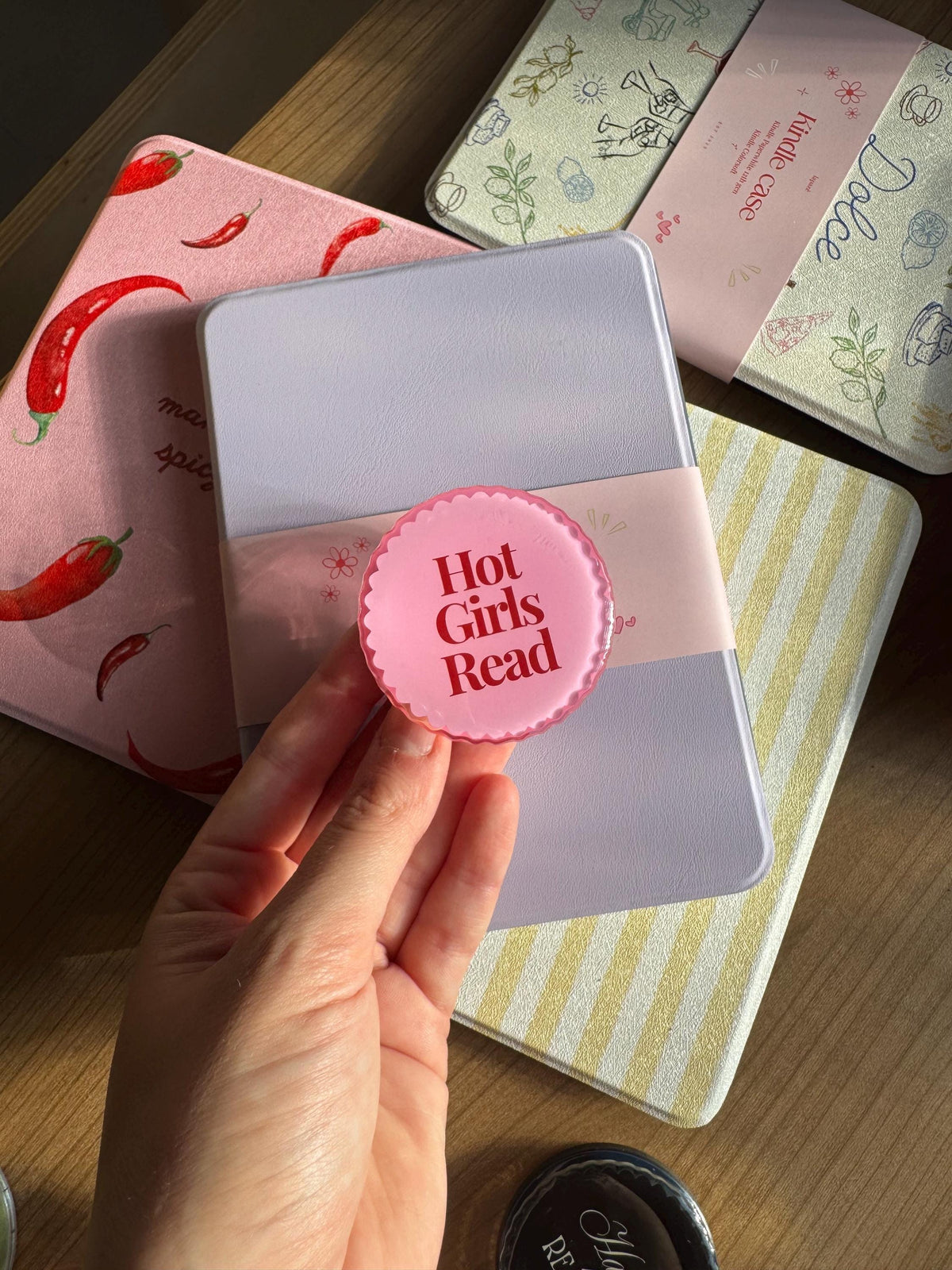 May include: A round pink scalloped-edge accessory featuring the text Hot Girls Read in dark red lettering, positioned over a light purple e-reader case with a matching pink paper band, surrounded by other e-reader cases with chilli and striped patterns.