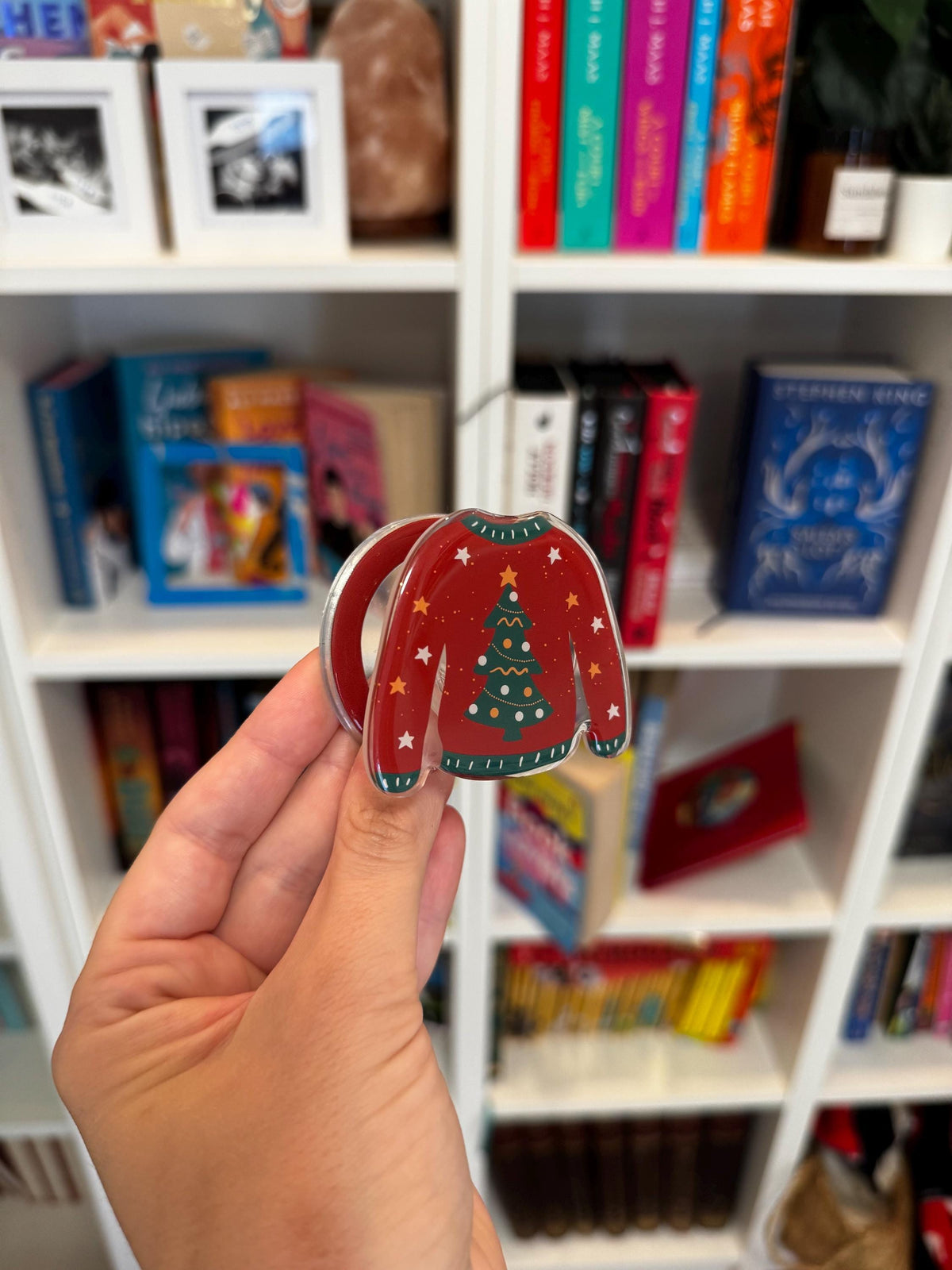 May include: A red phone grip shaped like a jumper featuring a green Christmas tree graphic with yellow star accents and white dots. The jumper design includes a dark green ribbed collar, cuffs, and hem with white dashed lines on the edges.