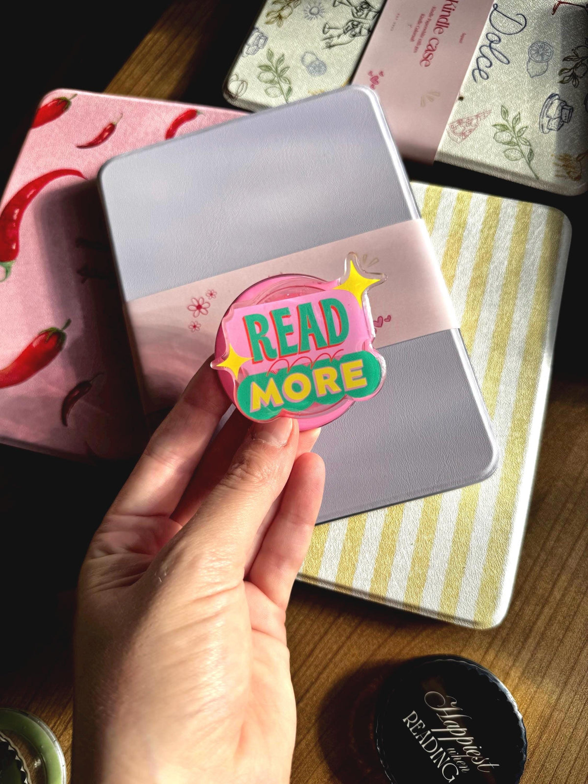 May include: A circular acrylic pin featuring a pink background with the text READ MORE in green letters, accented by two yellow four-pointed stars. The pin is held in front of a light purple Kindle case, which is surrounded by other patterned cases.