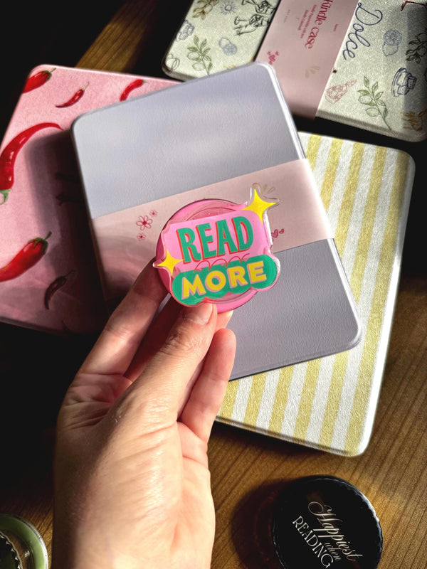 May include: A circular acrylic pin featuring a pink background with the text READ MORE in green letters, accented by two yellow four-pointed stars. The pin is held in front of a light purple Kindle case, which is surrounded by other patterned cases.