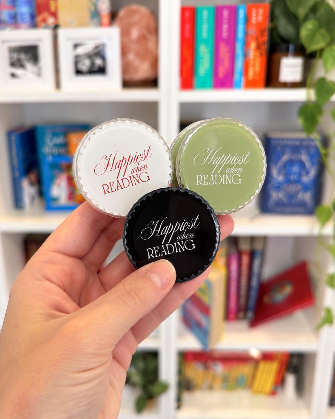 May include: Three circular jar lids with scalloped edges, each featuring the white or coloured text "Happiest when READING" in a script and serif font. One lid is white with red text, one is sage green with white text, and one is black with white text.