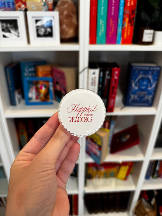 May include: A round, white, scalloped-edge pin featuring the red text "Happiest when READING" printed in the centre. The design has a smooth, glossy finish and a circular shape with decorative, wavy edges surrounding the central white surface.