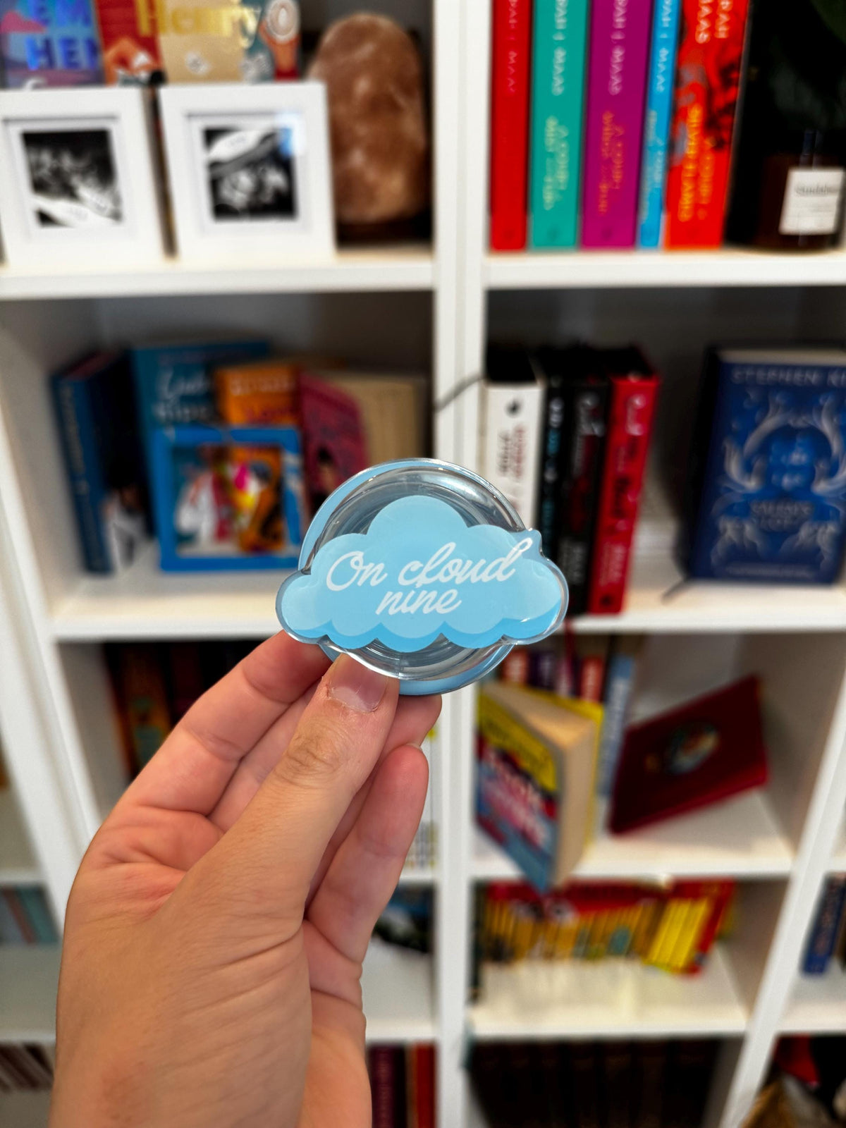 May include: A light blue phone grip shaped like a cloud with the white cursive text On cloud nine printed on the front. The accessory features a glossy finish and a circular base, designed to attach to the back of a mobile device for support.