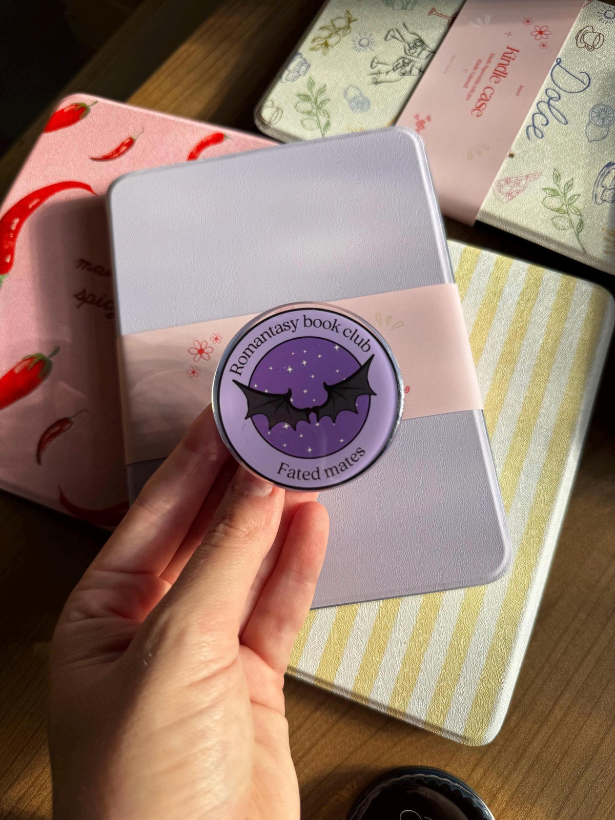 May include: A circular purple pin featuring a black bat wing graphic in the centre, surrounded by the text Romantasy book club and Fated mates. The pin is set against a light purple rectangular case wrapped in a pale pink paper band with floral accents.