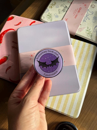 May include: A circular purple pin featuring a black bat wing graphic in the centre, surrounded by the text Romantasy book club and Fated mates. The pin is set against a light purple rectangular case wrapped in a pale pink paper band with floral accents.